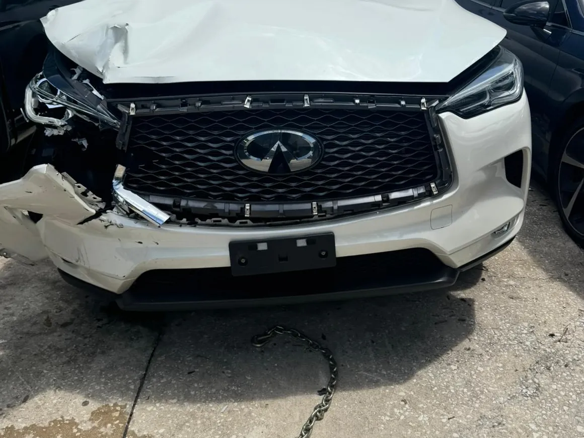 A white Infiniti QX50 on intake with the hood folded up, bumper cover torn off and the headlight hanging loose.