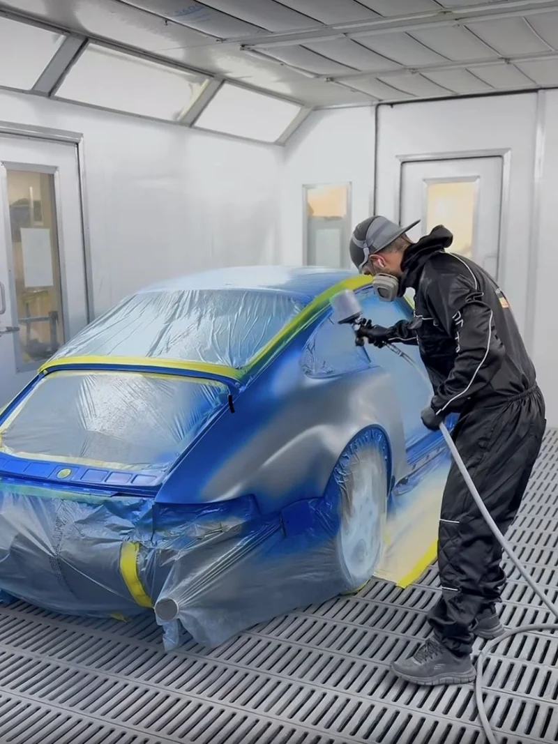 A painter in a full suit and respirator spraying blue base coat onto a masked Porsche 911 inside the paint booth.